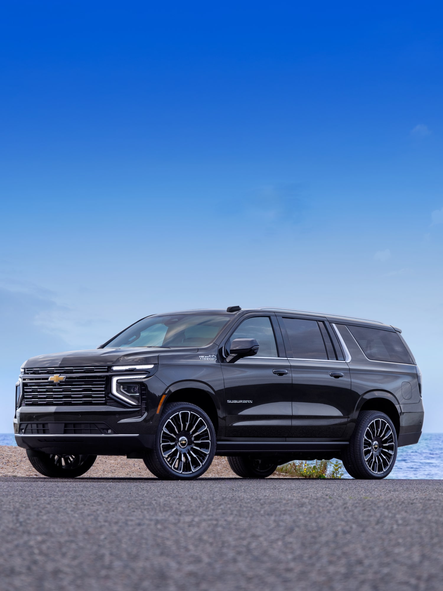 A Black Chevrolet Suburban Parked on a Rocky Shoreline With Blue Water and a Clear Sky in the Background.