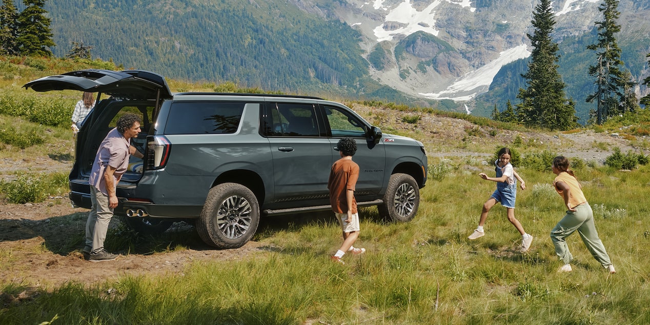 A Chevrolet Suburban Parked Beside a Mountain Meadow as Children Run and Play Near Tall Evergreen Trees.
