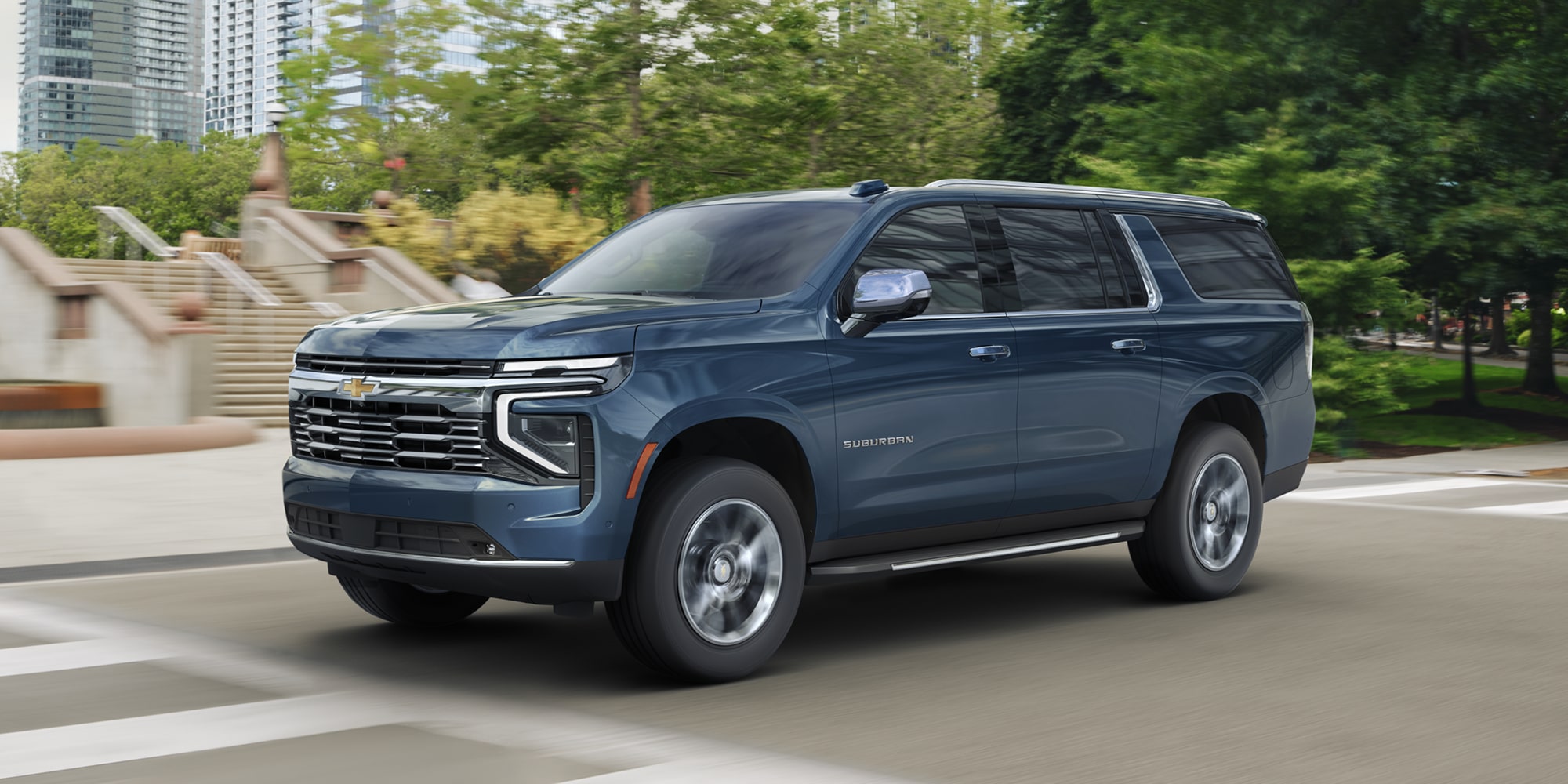 A Blue Chevrolet Suburban Driving Down a City Street With Green Trees and Stone Steps in the Background.