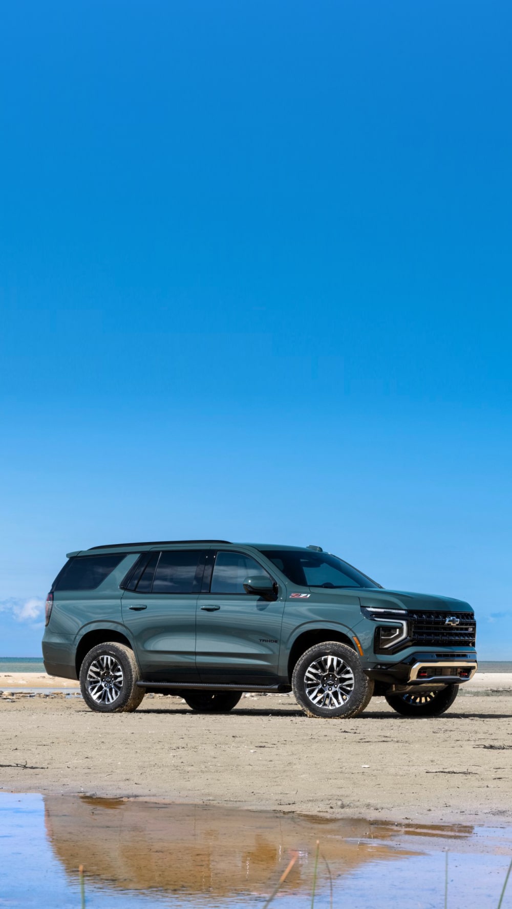 A Green Chevrolet Tahoe Parked on a Wide Sandy Beach Under a Bright Blue Sky With Light Clouds Along the Horizon.