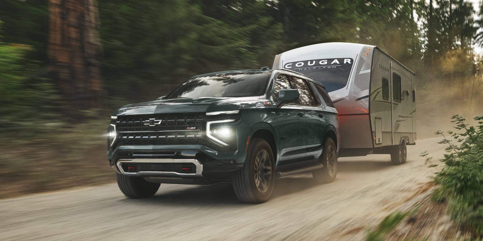 A Chevrolet Tahoe Towing a Large Silver Camper Trailer Down a Dusty Forest Road Surrounded by Tall Trees.