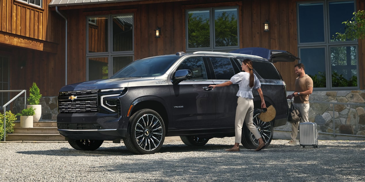 A Black Chevrolet Tahoe Parked in Front of a Modern Wood-Clad House as Two People Load Gear Into the Rear Seat.