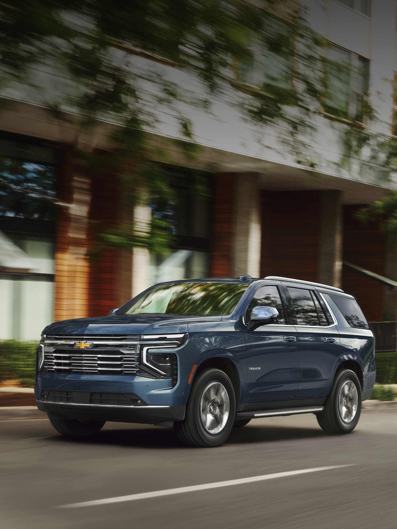 A Blue Chevrolet Tahoe Driving Down a Sunlit Urban Street Past Modern Brick Buildings