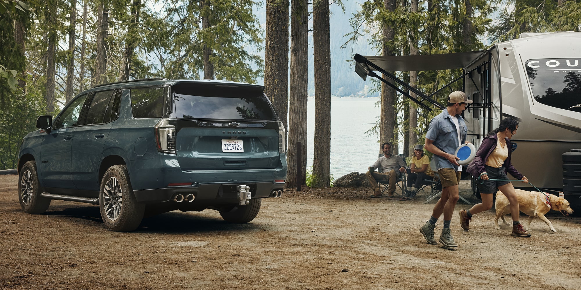 A Chevrolet Tahoe Parked Beside a Lakefront Campsite Where a Family Sets Up Gear and Plays With a Dog.