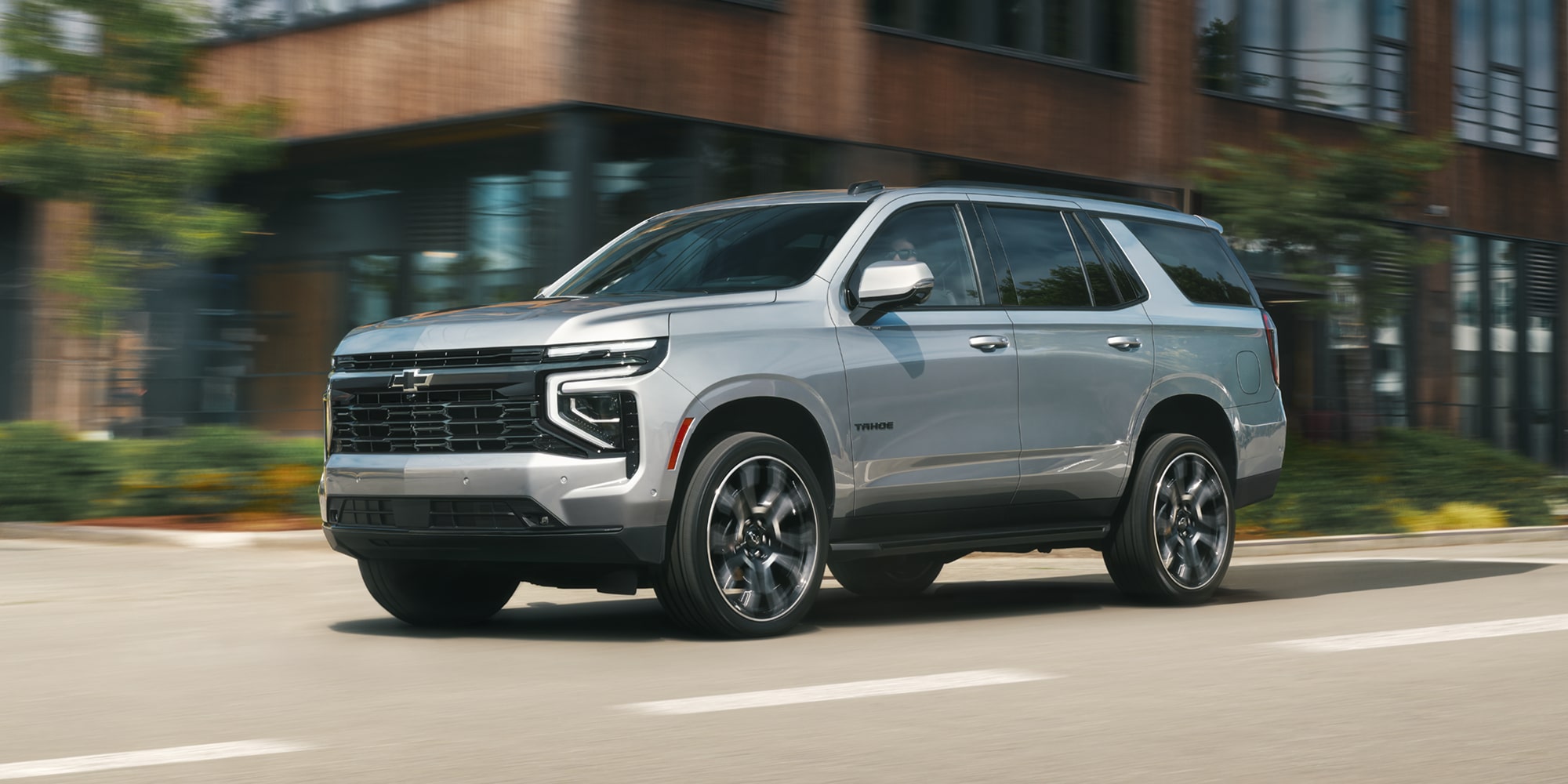 A Silver Chevrolet Tahoe Driving Down a Sunlit City Street in Front of a Modern Brick Building.