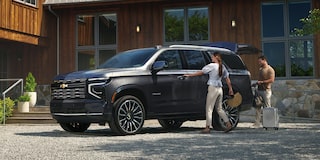 A Black Chevrolet Tahoe Parked in Front of a Modern Wood-Clad House as Two People Load Gear Into the Rear Seat.