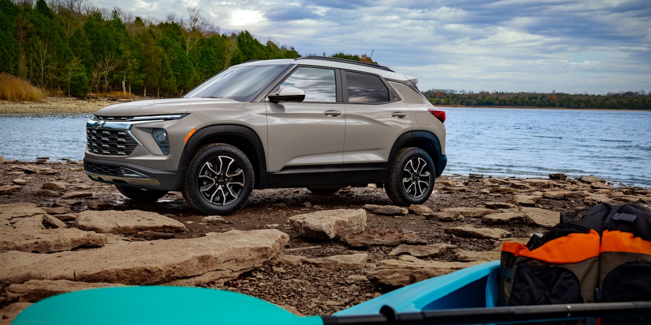A Chevrolet Trailblazer Parked on a Rocky Lakeside Shore next to Colorful Kayaks and Outdoor Gear under Bright Daylight.