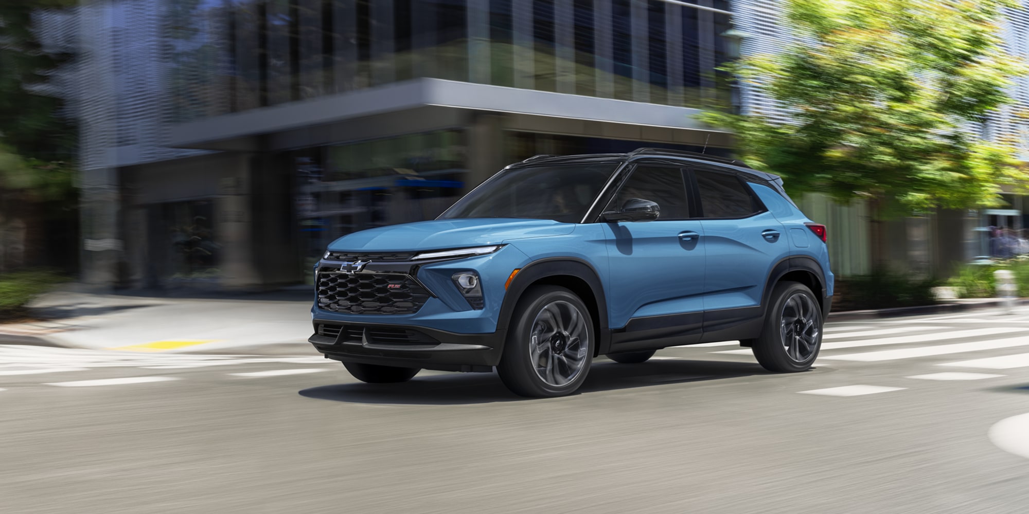 A Blue Chevrolet Trailblazer Driving Through a City Street with Modern Buildings and Green Trees in the Background.