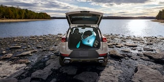 Una Chevrolet Trailblazer con la puerta trasera abierta que deja ver kayaks y equipos, estacionada en una orilla rocosa junto a un lago tranquilo.