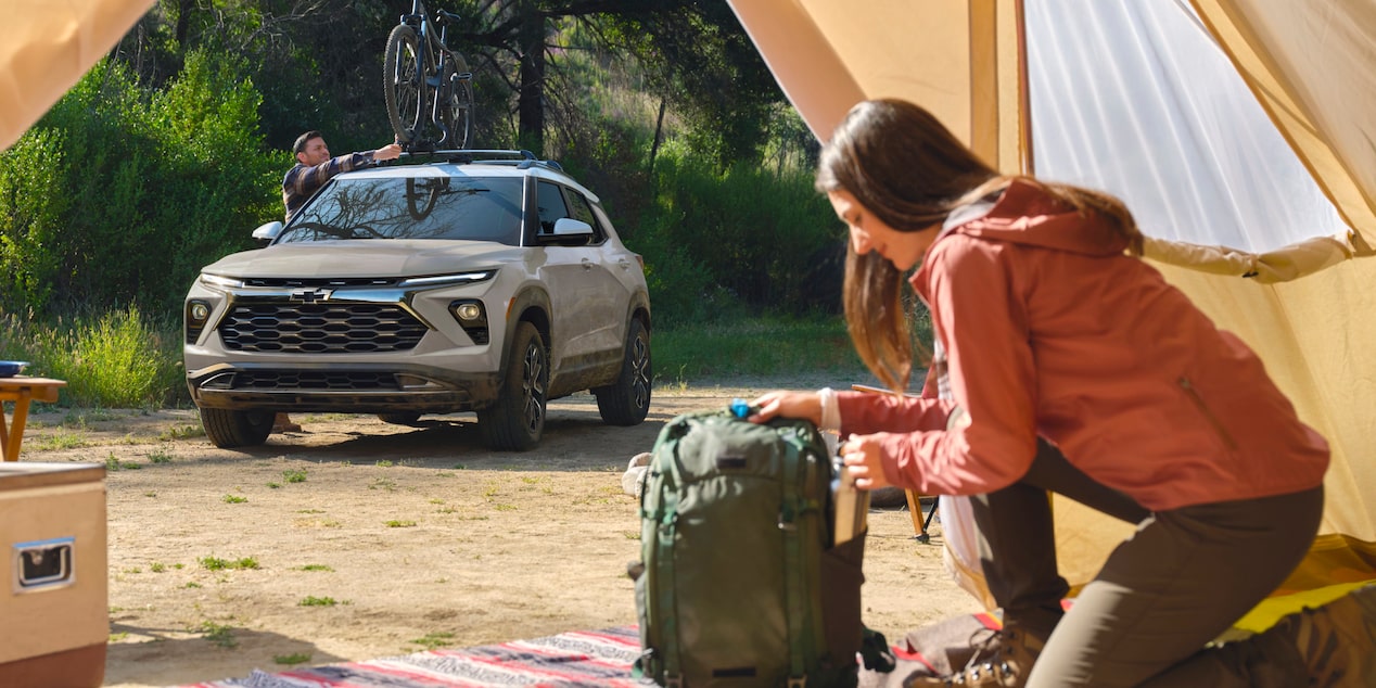 Una Chevrolet Trailblazer estacionada en un lugar de campamento mientras una persona carga equipos en una mochila junto a una carpa abierta.