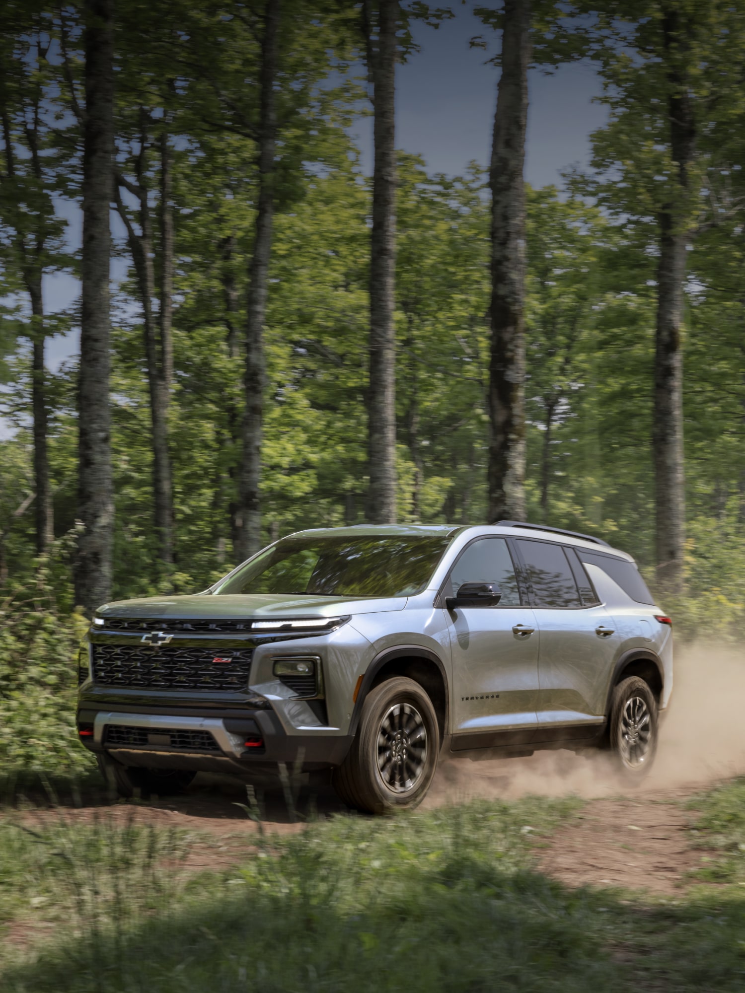 A Silver Chevy Traverse Driving Along a Dirt Trail in a Dense Forest While Kicking Up Dust.