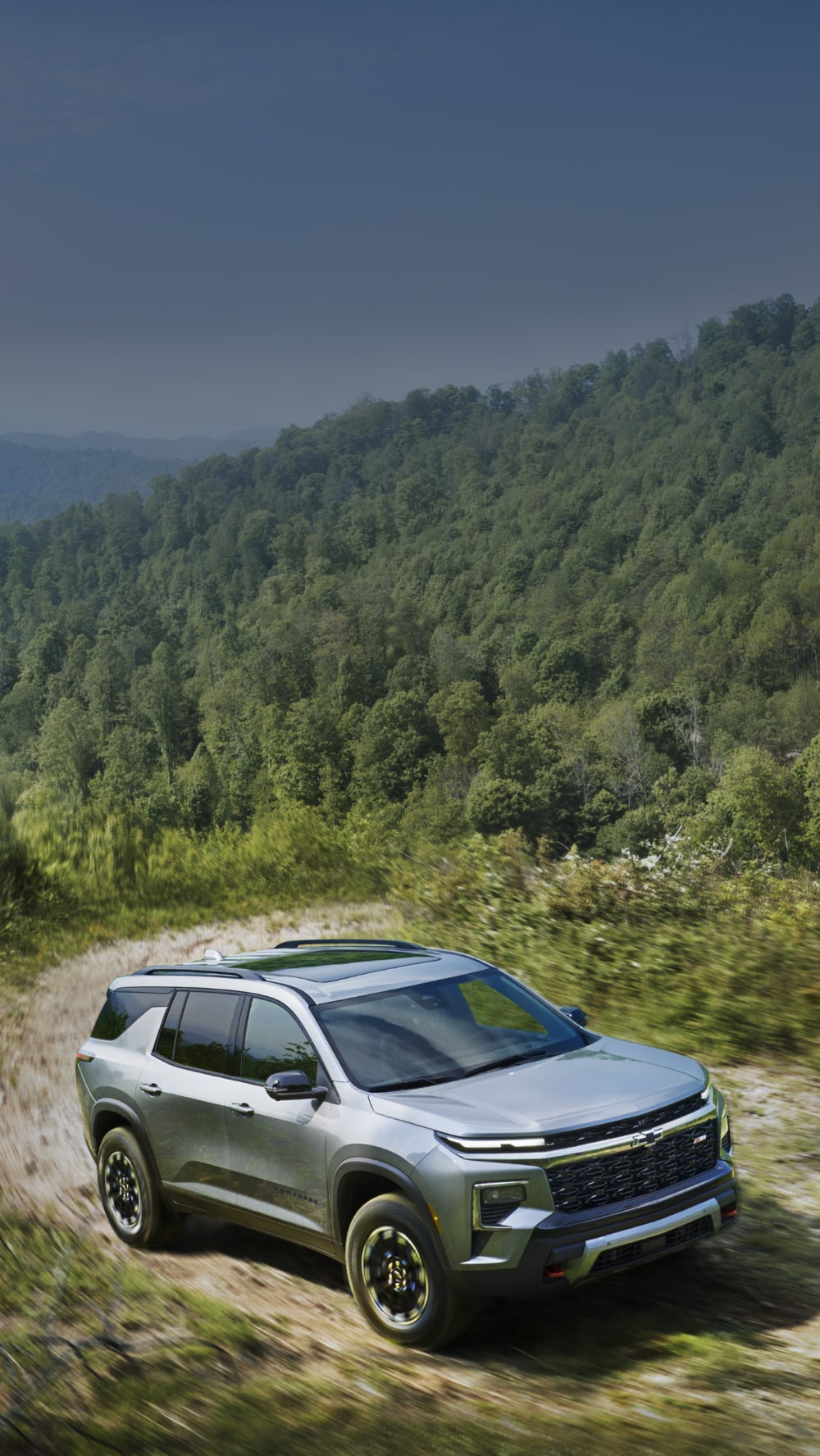 A Silver Chevy Traverse Driving Along a Dirt Trail in a Wide Open Landscape Surrounded by Dense Green Hills and Vegetation.