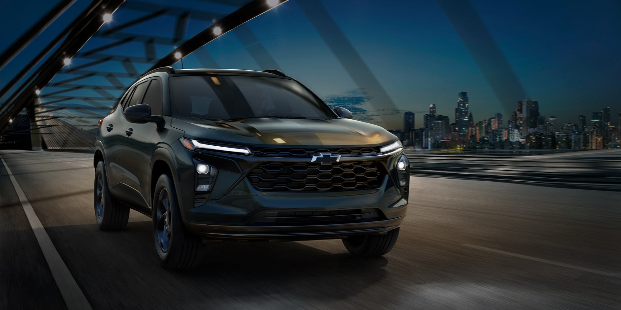 Chevrolet Trax cruzando un puente moderno al atardecer, con el perfil de la ciudad iluminado a lo lejos.