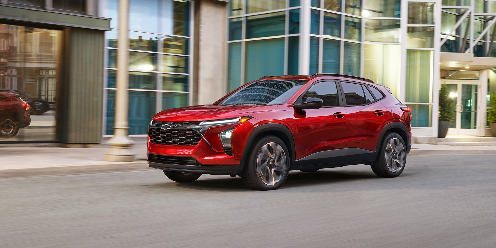 A Red Chevrolet Trax Driving Down a City Street in Front of Modern Glass Buildings.