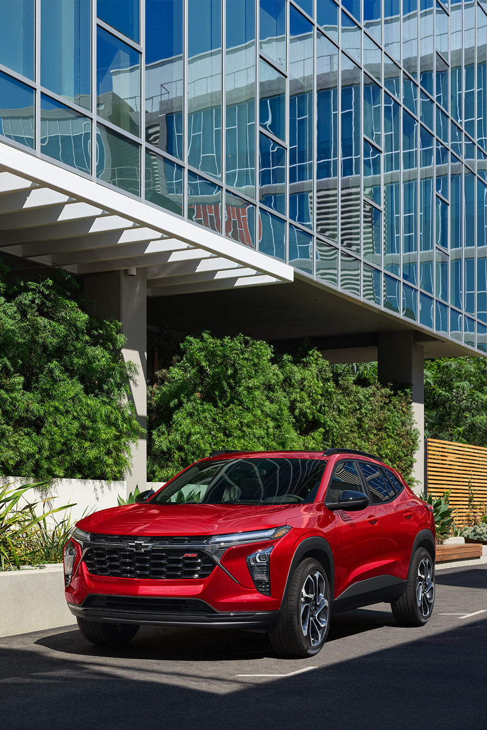 A Red Chevrolet Trax Driving Along a City Street With Sunlight Reflecting Off Its Front Fascia and Urban Landscaping in the Background