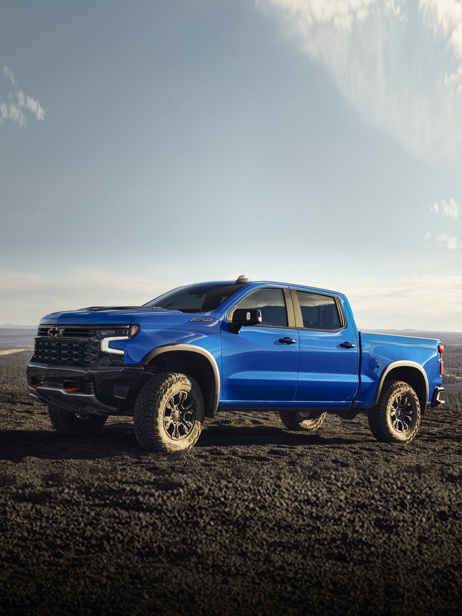 A Blue 2026 Chevy Silverado LD Pickup Truck Parked on Rocky Black Terrain Under a Bright Sky with Light Clouds.
