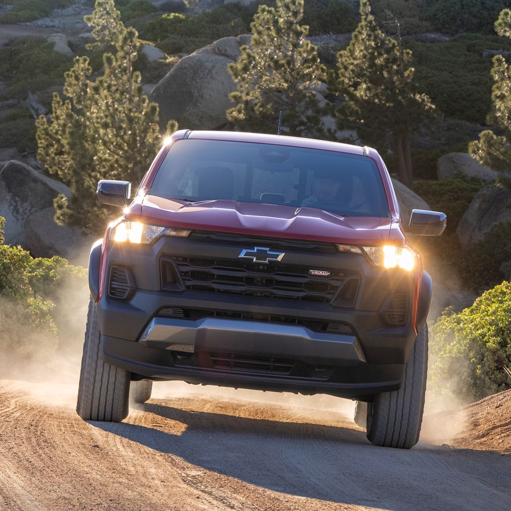 A Red Chevy Colorado Driving Along a Dirt Trail Through a With Headlights Illuminated and Dust Rising Behind the Truck.
