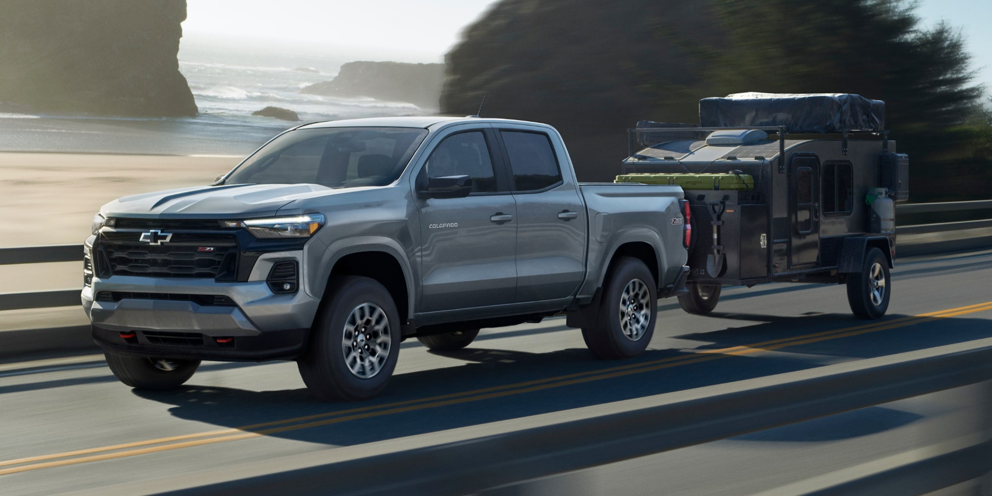 A Silver Chevy Colorado Towing an Enclosed Trailer Along a Coastal Highway With Ocean Cliffs in the Background.