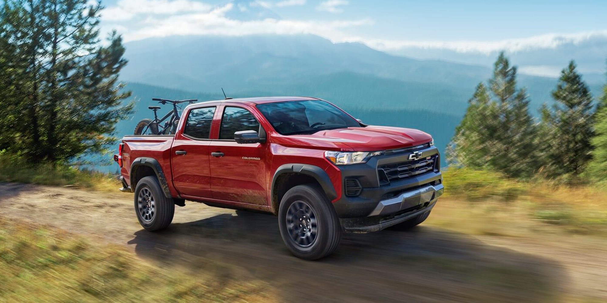 A Red Chevy Colorado Driving Along a Mountain Trail With Forested Hills and a Scenic Valley in the Background.
