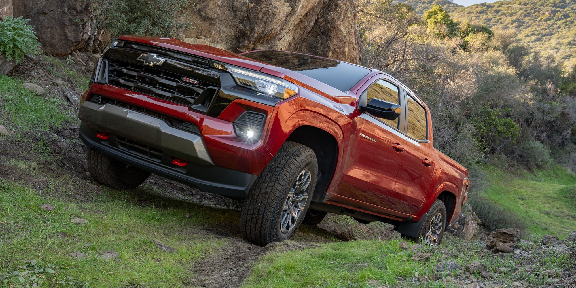 A Red Chevy Colorado Driving Along a Rugged Dirt Trail Surrounded by Green Vegetation and Rocky Hillsides.