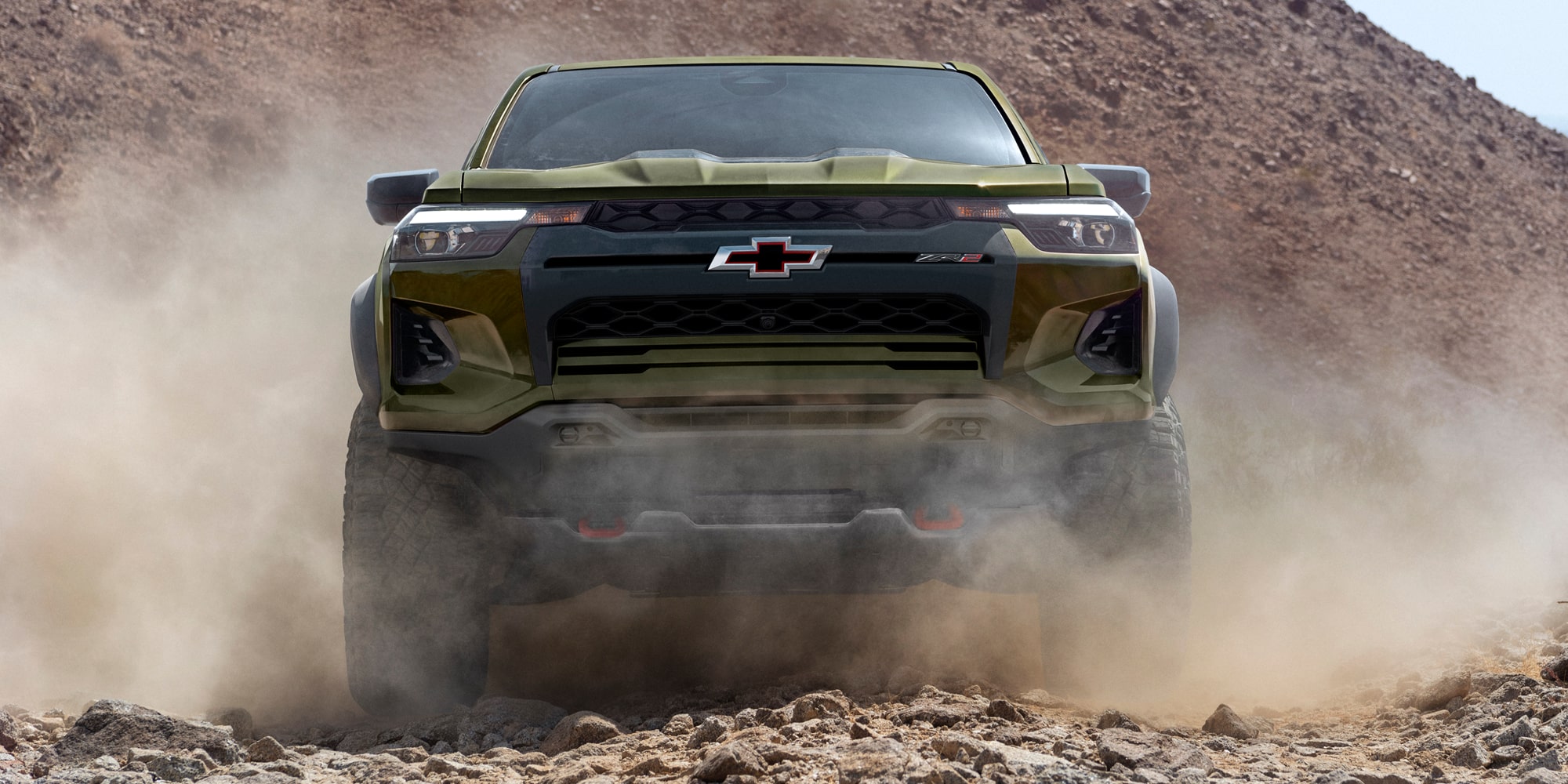 Front View of a Chevy Colorado Driving Through Dust on an Off-Road Trail With Headlights Illuminated.