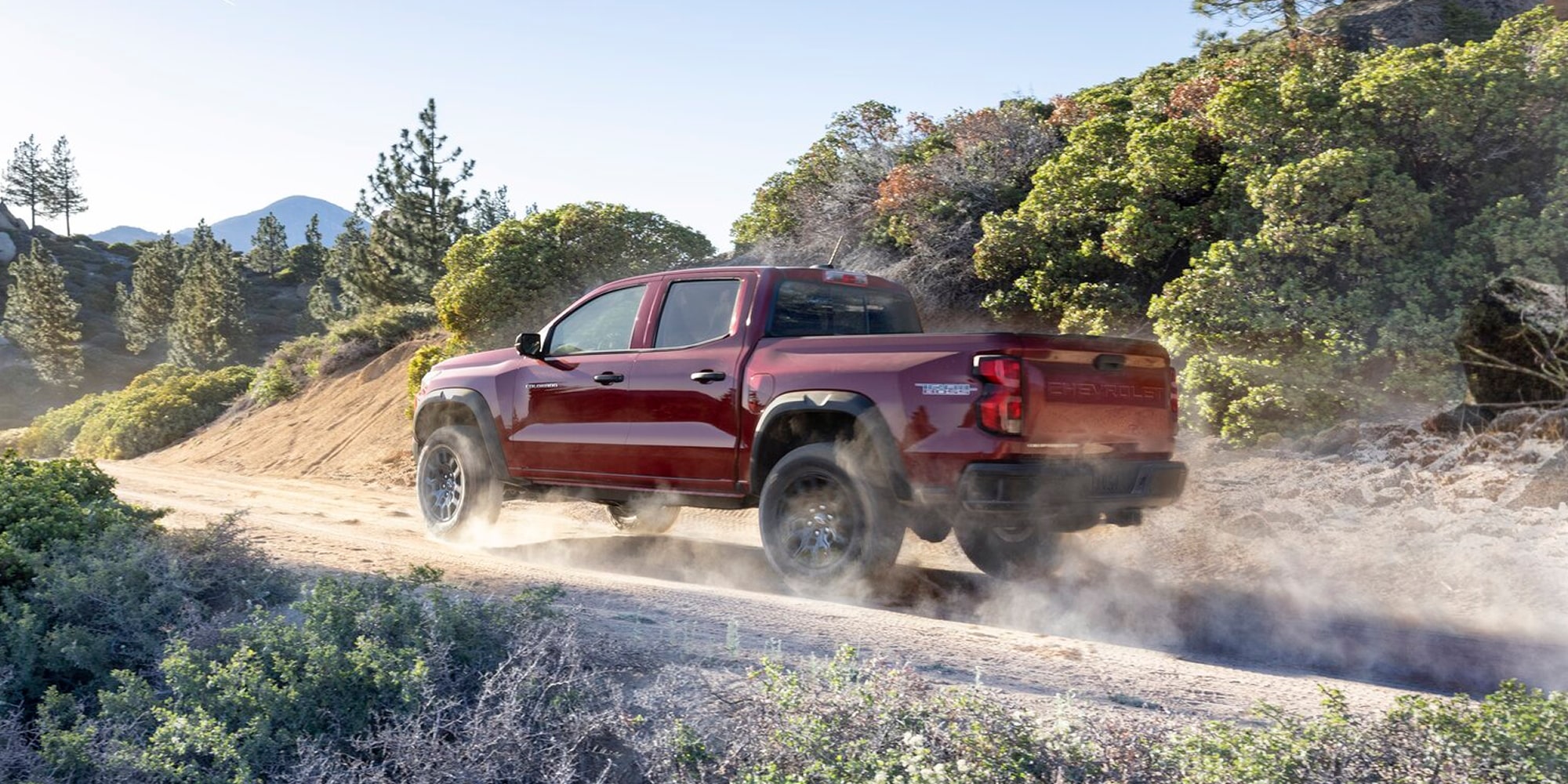 A Red Chevy Colorado Driving on a Dirt Road Through a Forested Landscape With Dust Trailing Behind the Truck.