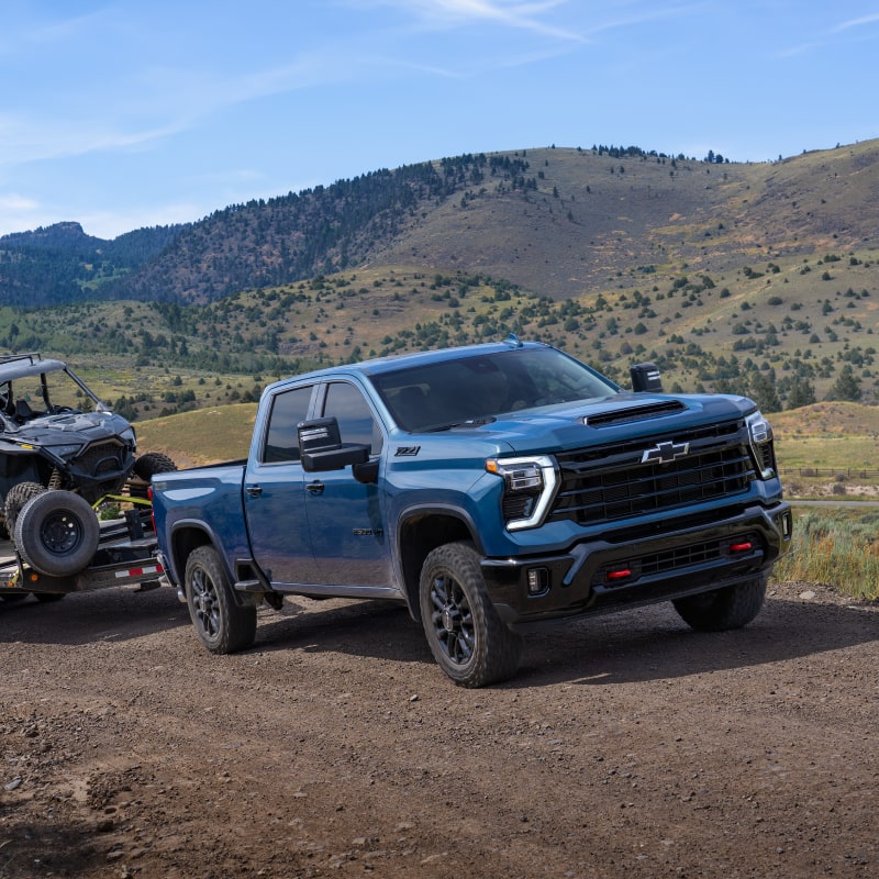 A Blue Chevy Silverado HD Towing an Off-Road Utility Vehicle Along a Mountain Road With Rolling Hills and Open Landscapes in the Background