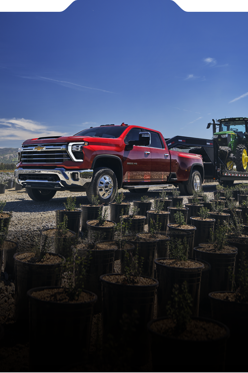 A Red Chevy Silverado HD Parked Among Cut Tree Stumps at a Worksite With a Green Tractor and Open Sky in the Background