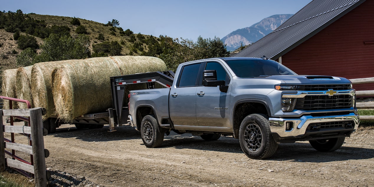 A Silver Chevy Silverado HD Towing a Large Hay Trailer Along a Rural Farm Road With a Red Barn and Mountain Landscape in the Background