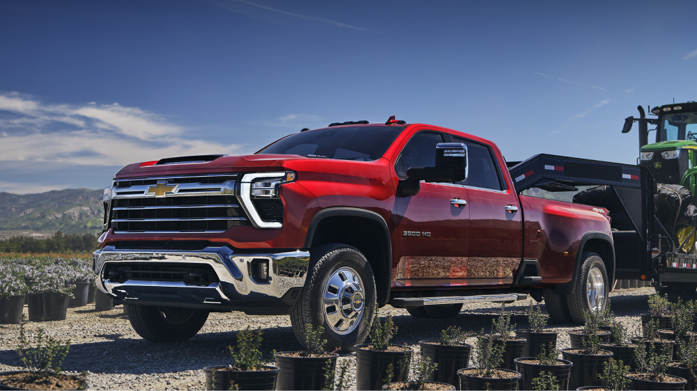 A Red Chevy Silverado HD Parked at a Logging Worksite Surrounded by Cut Tree Stumps With a Green Tractor and Mountain Landscape in the Background