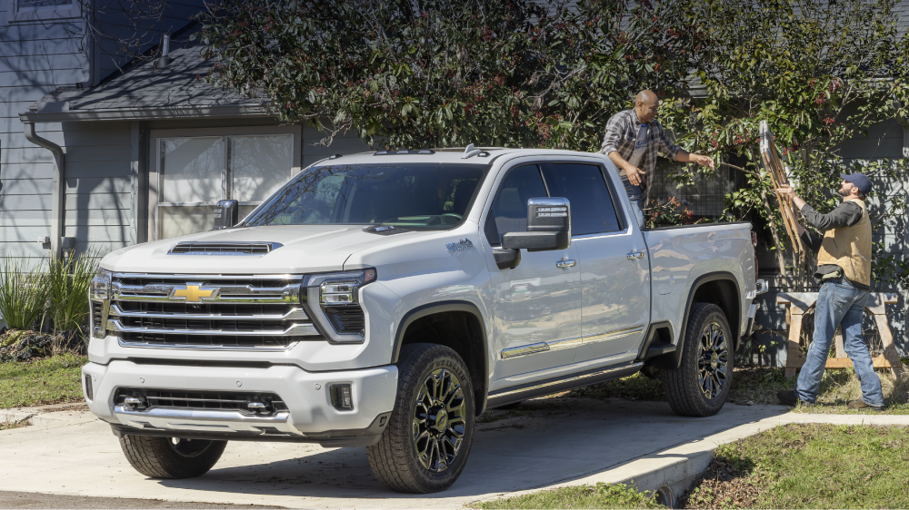 A White Chevy Silverado HD High Country Parked Outdoors With High Country Text Displayed Above the Truck and Landscaping Equipment Visible in the Background