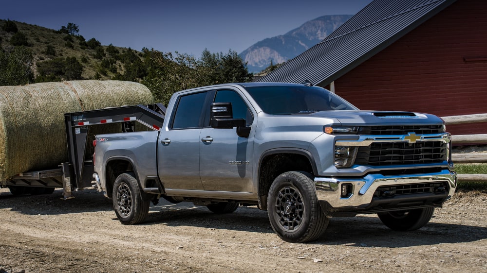 A Chevy Silverado HD Parked on a Dirt Road Near a Red Barn While Towing a Utility Trailer in a Rural Setting