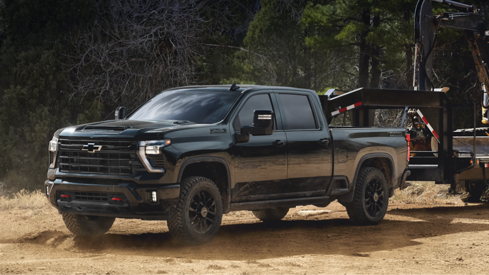 A Black Chevy Silverado HD Driving Across a Dusty Jobsite While Towing Equipment on a Trailer Through a Wooded Area