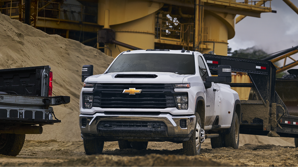 A White Chevy Silverado HD Driving Through an Industrial Worksite With Large Aggregate Piles, Heavy Equipment Structures, and Construction Vehicles in the Background