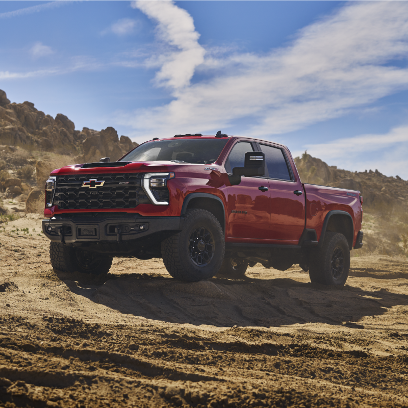 A Red Chevy Silverado HD Driving Across a Rugged Dirt Trail in a Desert Landscape With Rocky Mountains and Blue Sky in the Background