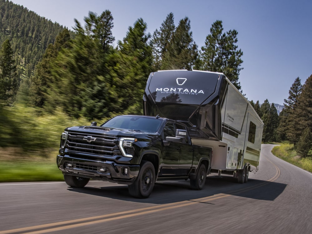 A Black Chevy Silverado HD Towing a Montana Fifth-Wheel Camper Through a Forested Mountain Road Surrounded by Tall Pine Trees