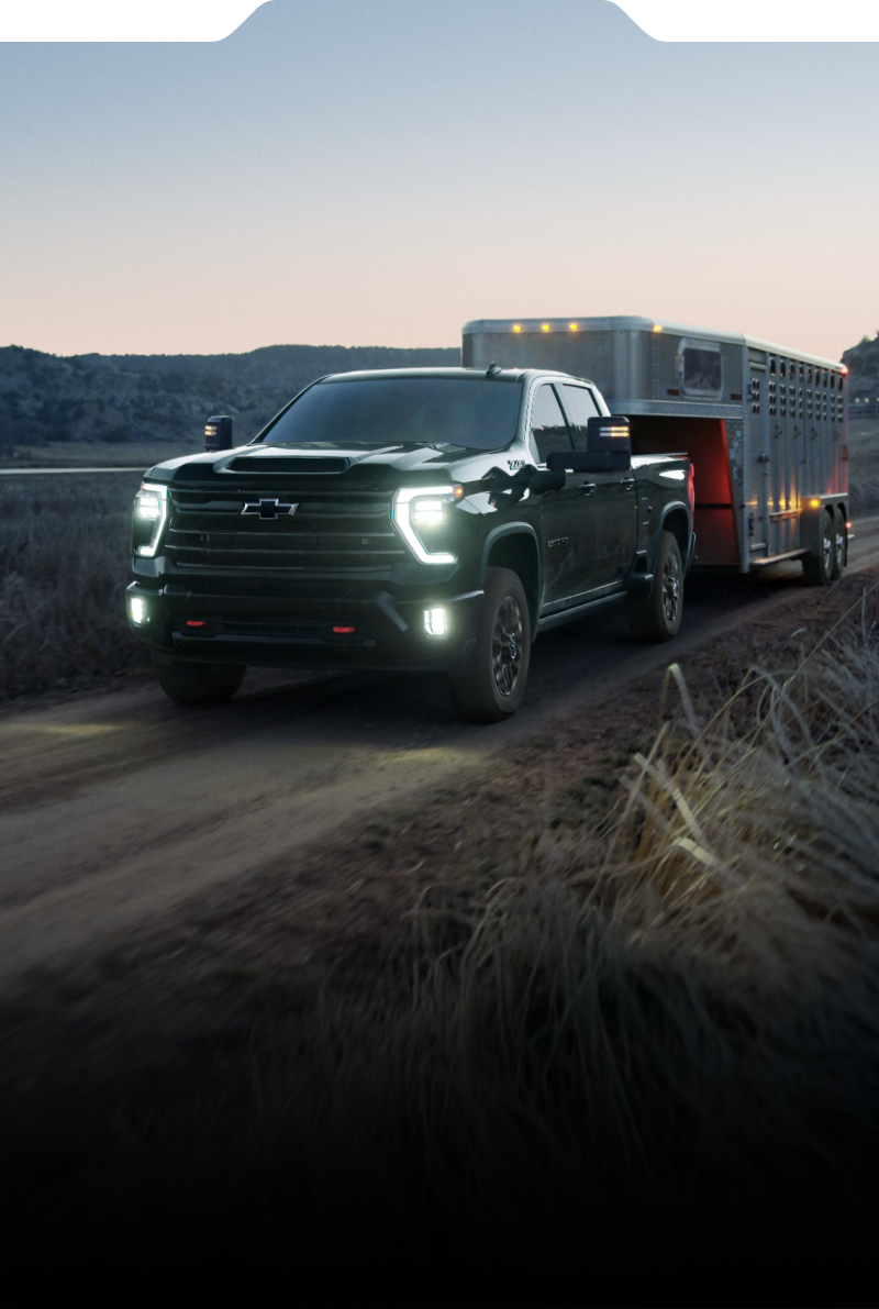 A Black Chevy Silverado HD Towing a Trailer Along a Rural Road at Dusk With Illuminated Headlights and Rocky Terrain in the Foreground