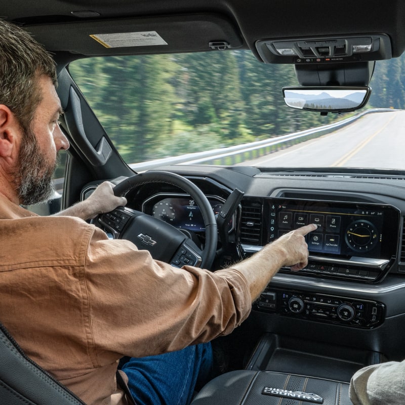 A Driver Using the Chevrolet Infotainment Touchscreen Inside a Chevy Silverado HD While Traveling on a Highway With the Road Visible Through the Windshield
