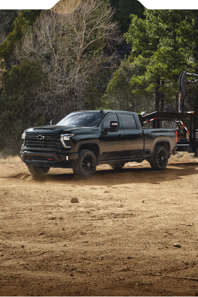A Black Chevy Silverado HD Towing a Flatbed Trailer Across a Dusty Worksite With Construction Equipment and Trees in the Background