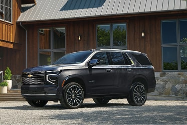 Side Profile View of a Black Chevrolet Suburban High Country Parked on a Forest Trail With Evergreen Trees and Snow-Capped Mountains in the Background.