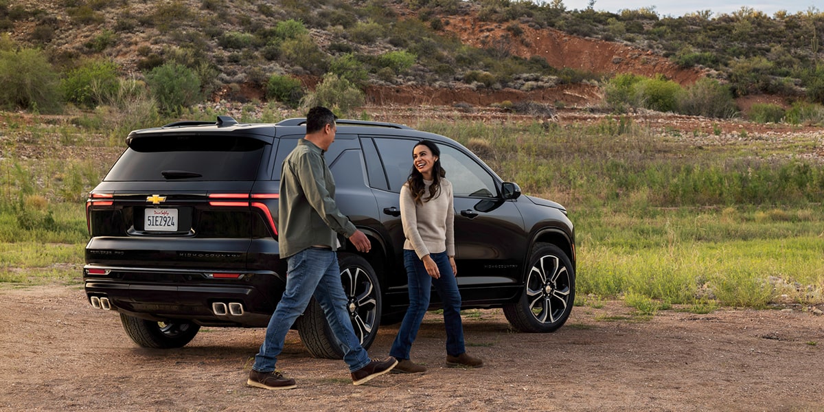 Chevrolet Traverse High Country Parked Outdoors With Two People Standing Beside the Rear Passenger Door and Rear Tailgate Area in a Natural Setting.