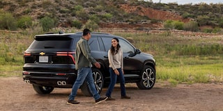 Chevrolet Traverse High Country Parked Outdoors With Two People Standing Beside the Rear Passenger Door and Rear Tailgate Area in a Natural Setting.