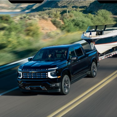 Chevrolet Silverado High Country Towing a Boat Trailer Along a Scenic Two-Lane Road With Rolling Hills in the Background.
