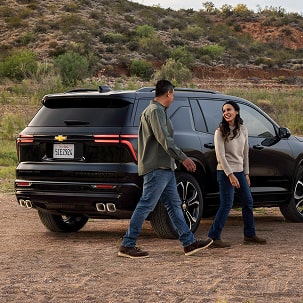 Chevrolet Traverse High Country Parked Outdoors With Two People Standing Beside the Rear Passenger Door and Rear Tailgate Area in a Natural Setting.