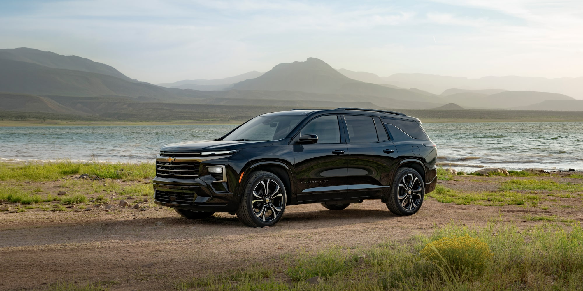 Front Three-Quarter View of a Black Chevrolet Traverse High Country Parked on a Dirt Road With a Lake and Mountain Peaks in the Background.