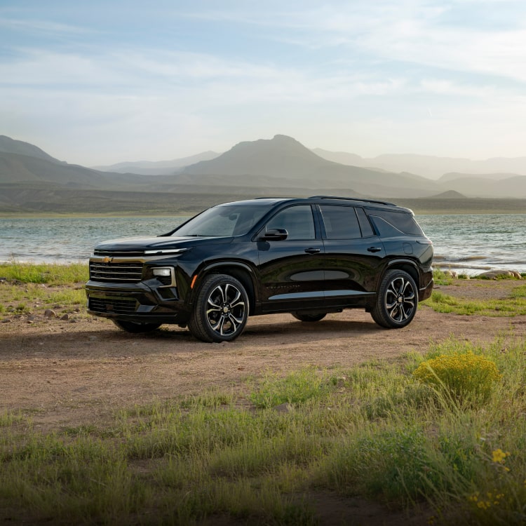Front Three-Quarter View of a Black Chevrolet Traverse High Country Parked on a Dirt Road With a Lake and Mountain Peaks in the Background.