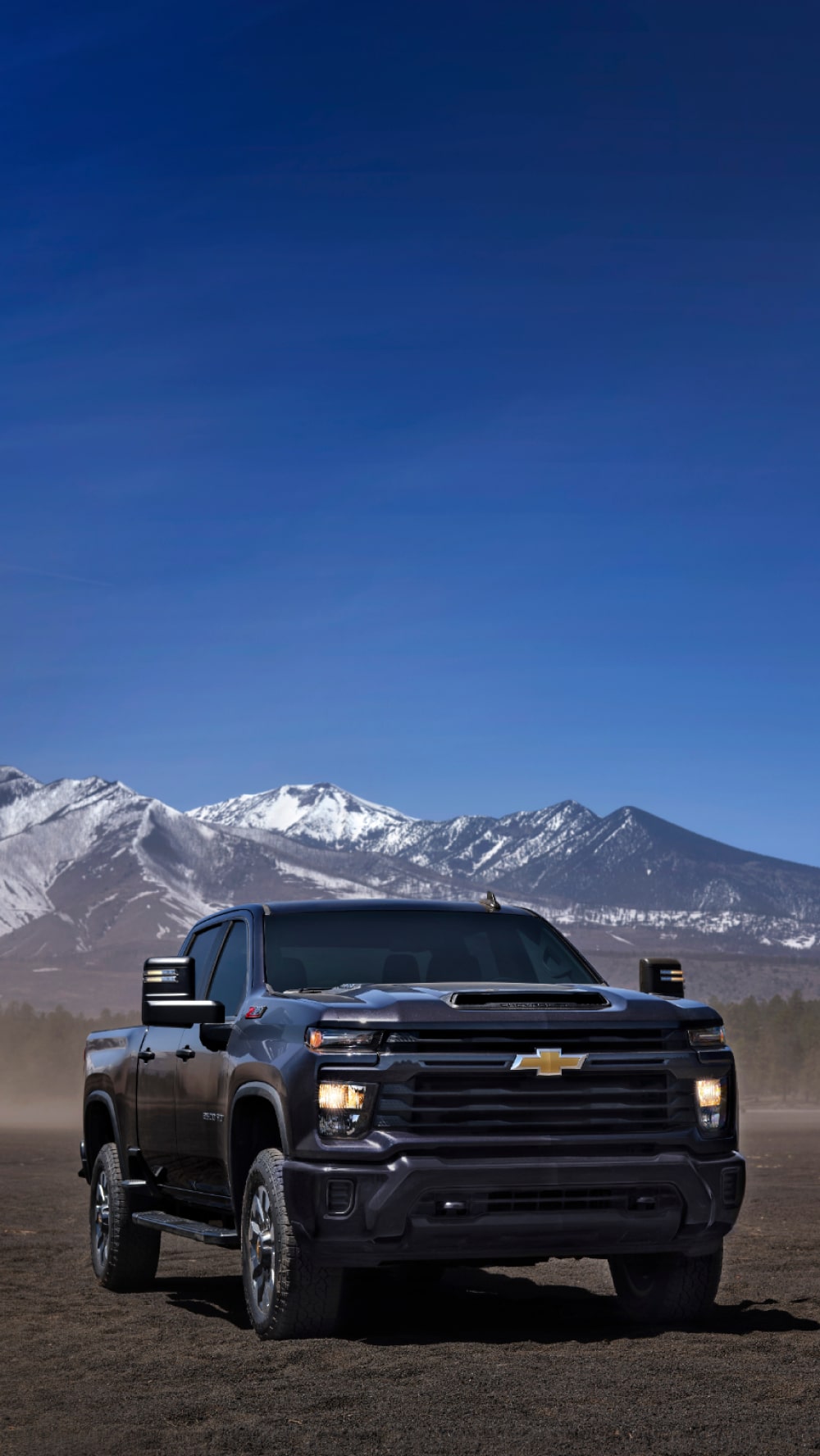 A Black Chevy Silverado HD Driving on a Dirt Road with Snow-Capped Mountains and Pine Trees in the Background Beneath a Clear Blue Sky.