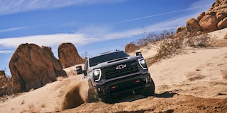A Black Chevy Silverado LD Climbing Over Large Desert Rocks with Dust and Sand Kicking Up from Its Tires.