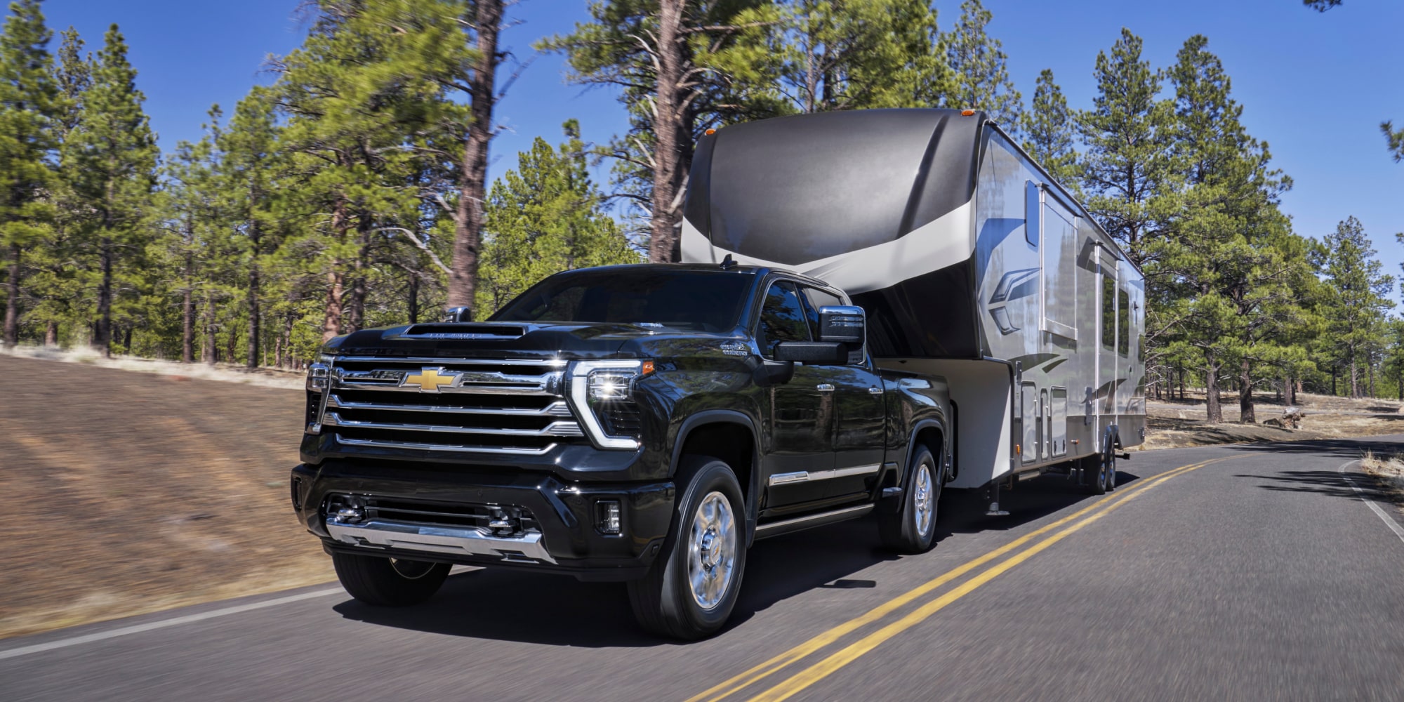 A Black Chevy Silverado LD Towing a Large Silver Camper Trailer on a Paved Road Lined with Trees and Mountains.