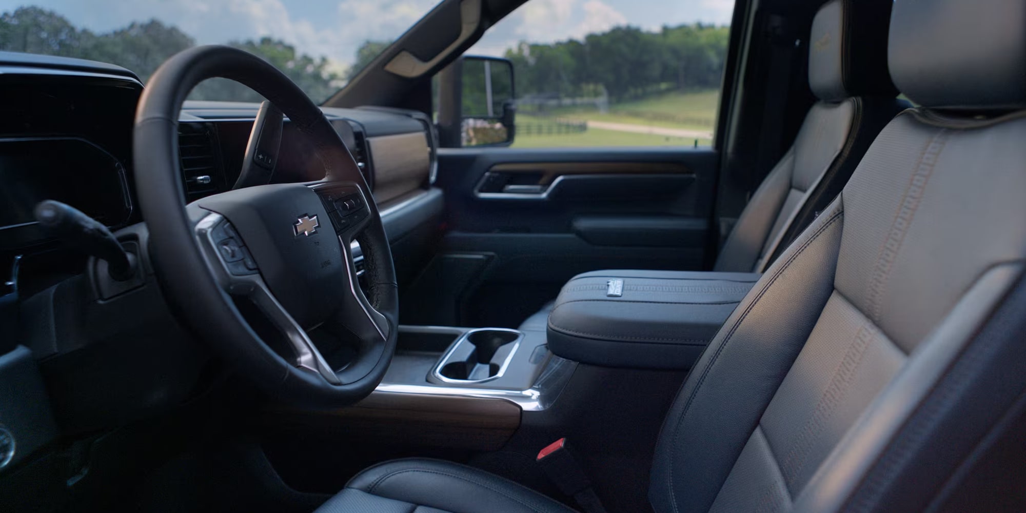 A Wide View Of The Chevrolet Silverado HD Interior Featuring Black And Gray Leather Seats, A Center Console With Cupholders, And A Steering Wheel Overlooking A Sunlit Rural Landscape Through The Windows.